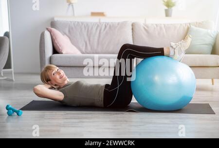 Fit mature lady doing abs exercises on yoga mat at home, training her core muscles with fitball, copy space Stock Photo