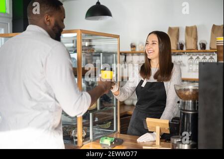Friendly and helpful waitress is serving an African-American male customer, preparing and giving paper cup of coffee to multiracial guy. Woman in apron holds out take-away coffee to guest of cafe Stock Photo
