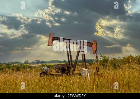 Old rusty oil well pump jack in field - home made modifications - with field and cows blurred in background and dramatic stormy evening sky Stock Photo