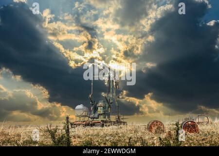 Oil well pump jack in field with old oil barrels behind barbed wire fence under dramatic sunset sky with dark oninous clouds Stock Photo