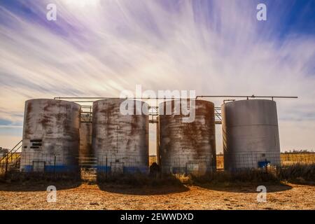 Old rusty holding tanks with shadows at oil well location on flat farm land under dramatic sky at golden hour Stock Photo