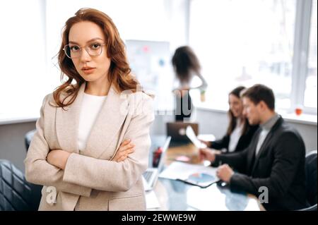 Creative modern office. Confident attractive redhead young woman, programmer or designer, stands in the office looks and smiles friendly at the camera, in the background colleagues are out of focus Stock Photo