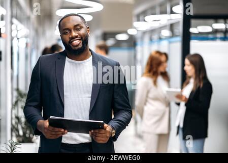 Creative modern office. Confident African American man, ceo, programmer or designer, stands in the office looks and smiles friendly at the camera, in the background colleagues are out of focus Stock Photo