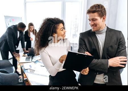 Two multiracial successful colleagues communicate in a modern office, discuss new ideas, think about a plan, in the background their colleagues Stock Photo