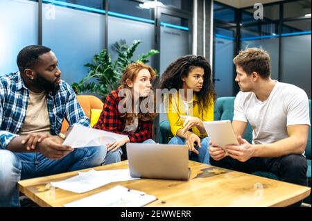 Concentrated coworkers or students sitting on the couch, discussing work or study project. Serious multiethnic colleagues working together on presentation or financial report Stock Photo