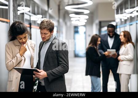 Concept of office work. Concentrated business partners standing in hall, discussing corporate project. Multiracial young adult colleagues talking about some company plans, giving each other advices Stock Photo