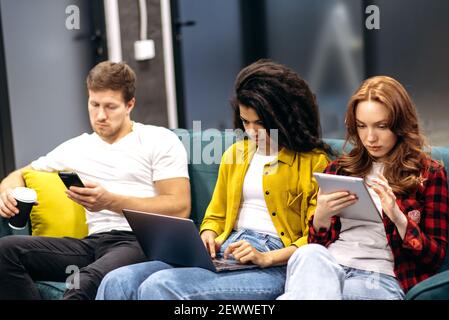 Focused young adult coworkers sitting on the couch in office, using gadgets, working together. Concentrated multiracial employees searching ideas for corporate project, teamwork concept Stock Photo