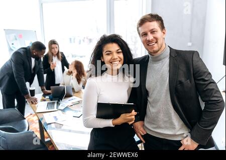 Portrait of two multiracial successful colleagues, African American female and caucasian male, standing in the modern office, looking and smiling at the camera Stock Photo