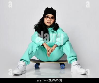 Young asian girl in black hat and glasses in pastel modern green, mint color hoodie and pants sitting on skateboard on white