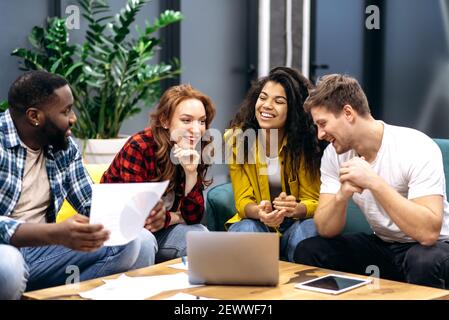Friendly multiracial business team, sitting in office, talking about work process, smiling, communicate with each other, discuss about new startup, studying financial graphs Stock Photo