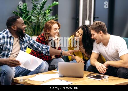 Joyful friendly employees or students take a break from work or study, sitting on the sofa, telling funny stories. Multiracial male and employee coworkers are having a pleasant conversation, laughing Stock Photo