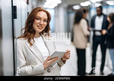 Creative modern office. Confident attractive redhead young woman, programmer or designer, stands in the office looks and smiles friendly at the camera, in the background colleagues are out of focus Stock Photo
