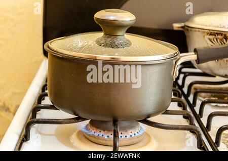The flame of the stove burner rises under a black non-stick pot  that is covered with a glass lid with water droplets on the inside. Stock Photo