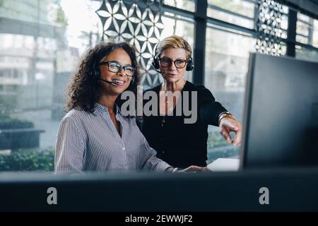 Two businesswomen cooperating while using computer and working in the office. Manager helping female colleague working on computer. Both wearing heads Stock Photo