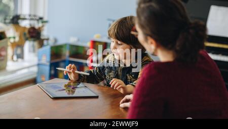 Kid using digital tablet with his mom sitting in front around the table. Boy learning to draw on graphic tablet with mother sitting by. Stock Photo