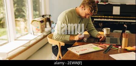 Freelance illustrator working from home office. Man sitting at table with laptop making drawings on his graphic tablet using stylus pen. Stock Photo