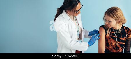 Female doctor giving vaccine to young woman in clinic. Healthcare worker putting bandage on arm of a woman after giving vaccination. Stock Photo