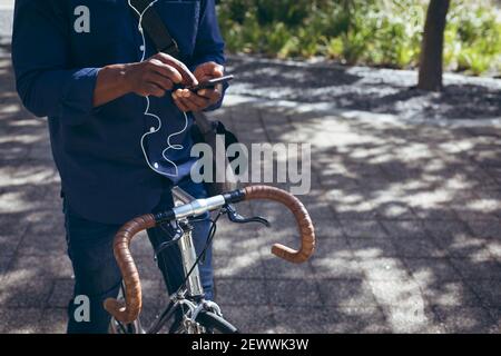 Midsection of african american senior man wearing earphones sitting on bicycle in street using smart Stock Photo