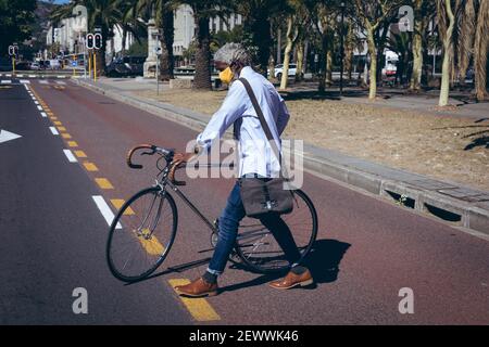 African american senior man wearing face mask wheeling bicycle across road on a pedestrian crossing Stock Photo