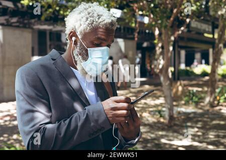 African american senior man wearing face mask and earphones standing in street using smartphone Stock Photo