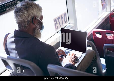 African american senior man wearing face mask sitting on bus using laptop Stock Photo