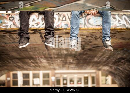 Two friends with legs hanging off concrete roof in skatepark Stock Photo