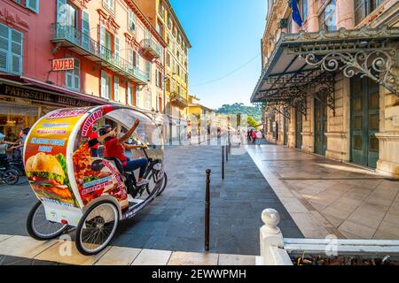 A female cyclist taxi driver guides tourists on a 3 wheel rickshaw bicycle through the Old City of Nice, France on the French Riviera.