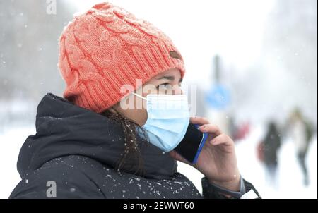 The picture shows a Caucasian woman with cap and mask for the coronavirus talking on her smartphone during a snowfall Stock Photo