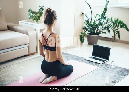 Back view photo of a brunette woman sitting in front of the laptop doing fitness exercises on the floor Stock Photo