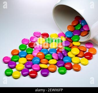 A white mug pouring colorful chocolate buttons on a white background. Stock Photo