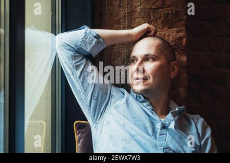 Pensive man in a blue shirt looking out the window with sadness and thinking. Stock Photo