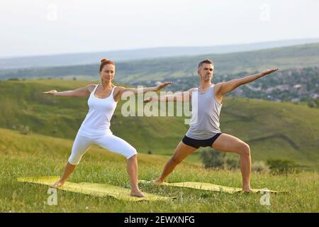 Middle aged couple doing Warrior II pose while practicing yoga on grassy hill in countryside Stock Photo