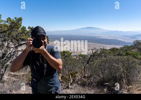 A bearded male photographer holding his camera and standing on the hill against the mountains Stock Photo