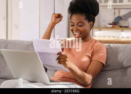 Excited Black woman sit on sofa at home receiving job enjoy exam results or college admission letter Stock Photo