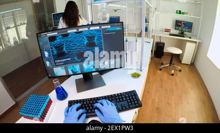 POV of researcher doctor typing at computer, analysing DNA scan image, while coworker bringing him samples of blood. Group of biochemistry scientists examining virus evolution using high tech Stock Photo