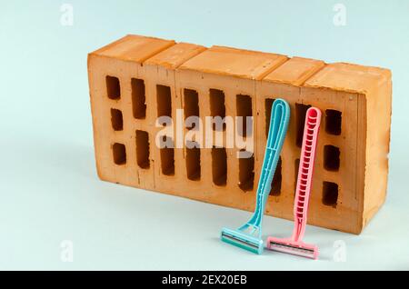 Two plastic razors and a red brick on a blue background. Blue and pink razors are leaning against a brick with holes. Selective focus. Stock Photo