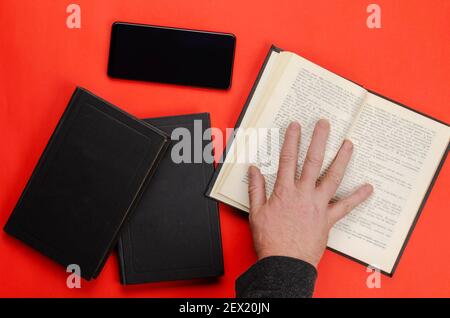 A man's hand holds an open paper book on a red table. A black smartphone and two closed books lie side by side.  Flat lay Stock Photo