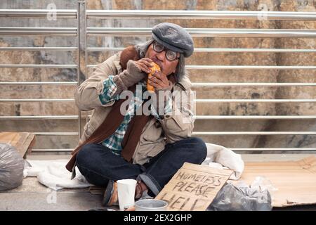 Aged dirty homeless begger eating bread on the bridge Stock Photo