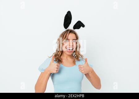 Happy blonde young woman with easter bunny ears smiling showing thumbs up gesture while standing on white studio background. Stock Photo