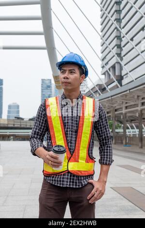 Young asian constructor with safety helmet in work vest holding coffee cup in construction site Stock Photo