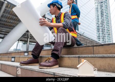 Wooden toy house on constructor team sitting with layout paper and survey on construction site in the city Stock Photo