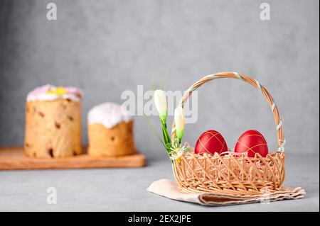 Two red painted Easter eggs in the wicker basket on gray table top with two easter cakes on backdrop Stock Photo