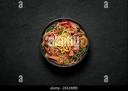 Japchae in black bowl on dark slate table top. Korean cuisine glass chapchae noodles dish with vegetables and meat. Asian traditional food. Authentic Stock Photo