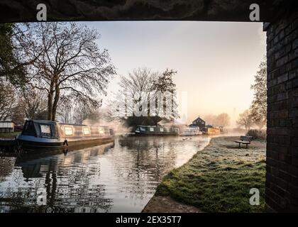 Stunning old canal house boats landscape under bridge sunrise in countryside with river and single lone wooden bench. Frost on grass reflection trees Stock Photo