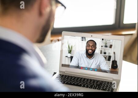 Back rear view focused young African male employee working from home using computer application, involved in video conference brainstorming meeting with male team leader, planning a new strategy Stock Photo