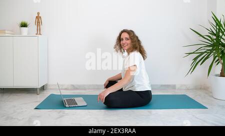 Young woman with curly blond hair practicing a yoga class with her computer from home. She has a blue mat on the marble floor. Stock Photo