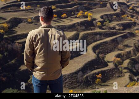 Guy in shirt and jeans looks at large terraced hills with brown fields and orange yellow trees under bright sunlight on autumn day Stock Photo
