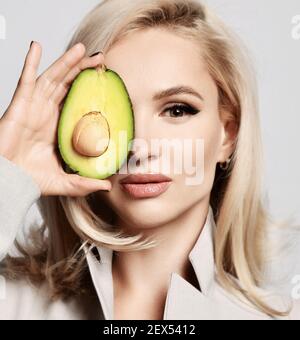 Pretty woman with blonde wavy hair and healthy skin wearing beige shirt holds half an avocado covering half of face Stock Photo