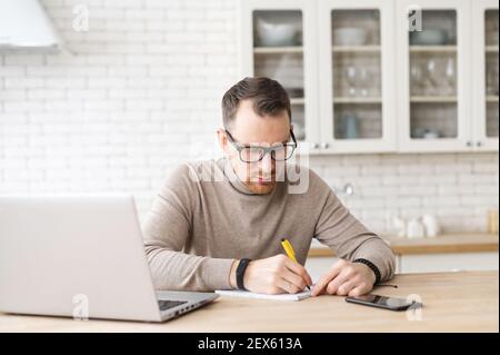 Serious intelligent concentrated young businessman freelancer working on laptop, doing online market research, working on project from home, creating plan or checking financial statement, taking notes Stock Photo