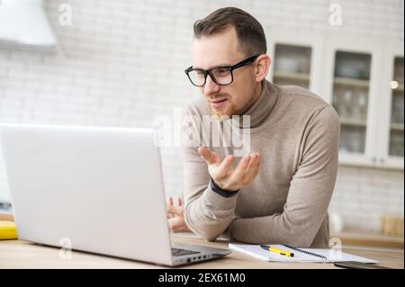 Excited young adult bearded hipster man in glasses land casual clothes leaning on the kitchen table and looking at laptop screen, video calling online, chatting having discussion with a friend Stock Photo